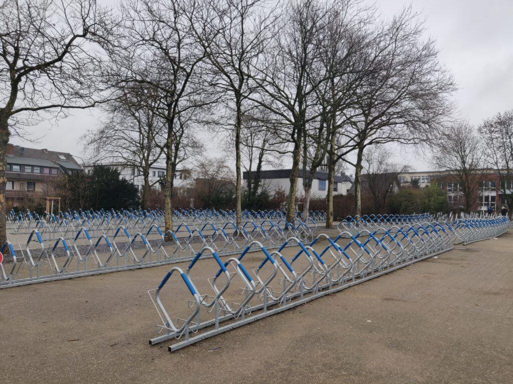 Reihenweise Fahrradparker aus Metall auf einem großen, offenen Platz unter Bäumen