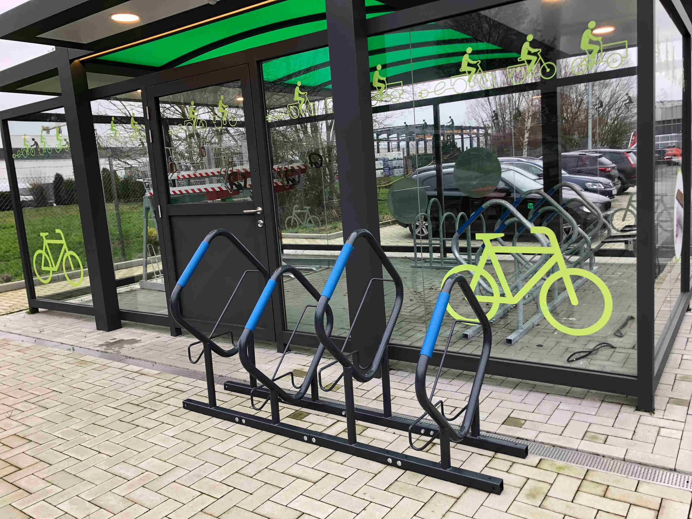 Moderner Fahrradparker aus Metall vor verglaster Fahrradüberdachung mit Fahrradmotiven