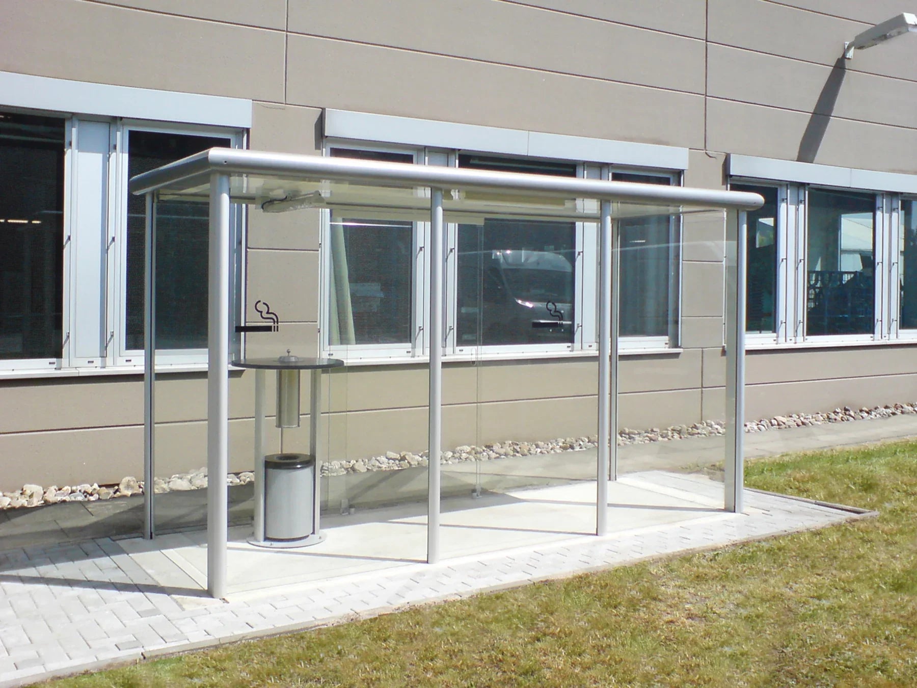 Raucherunterstand aus Metall mit Glaswänden und Dach, auf Pflaster vor Firmengebäude