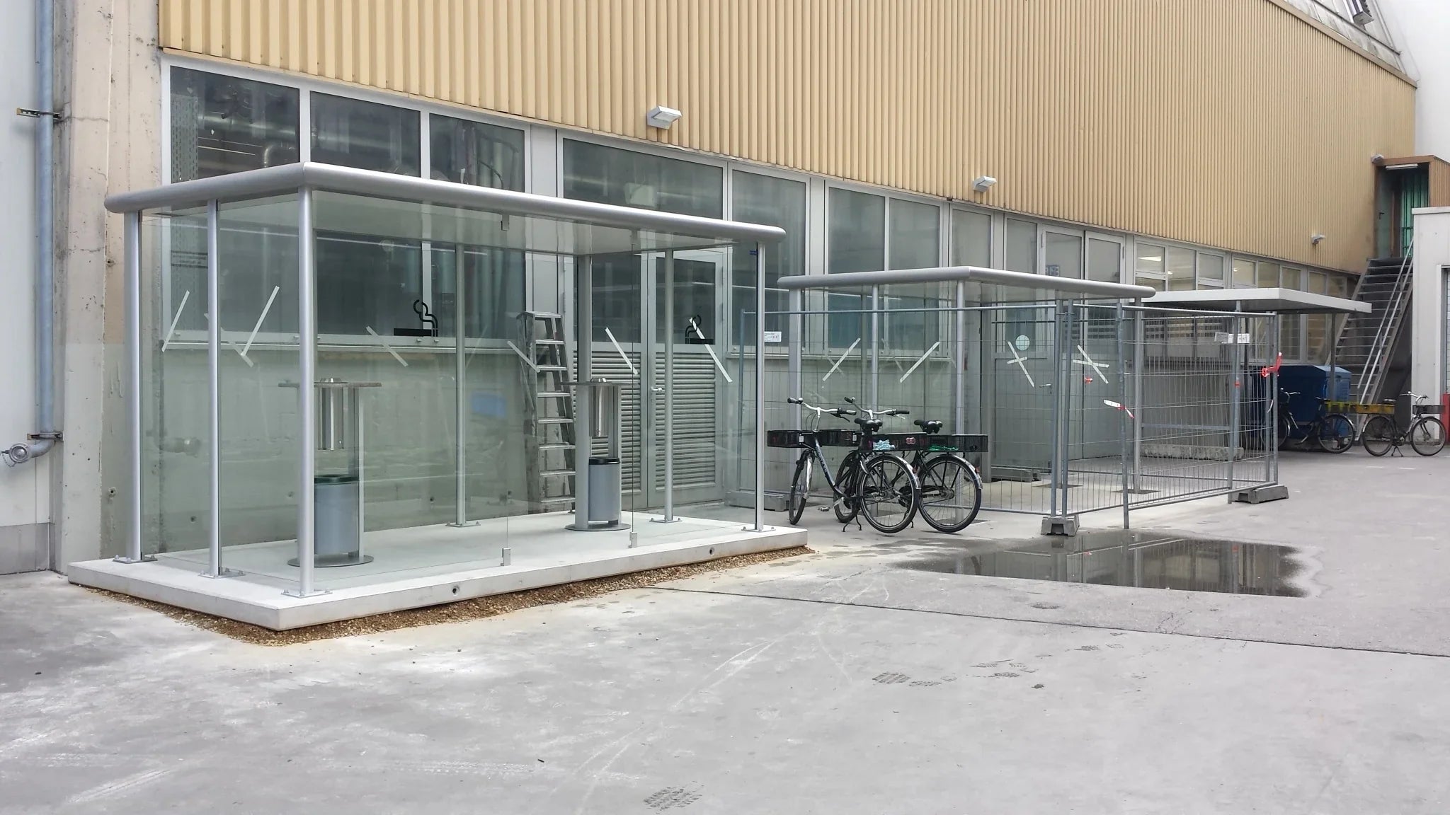 Überdachte Raucherunterstände und Fahrradständer aus Metall und Glas auf einem Firmenhof