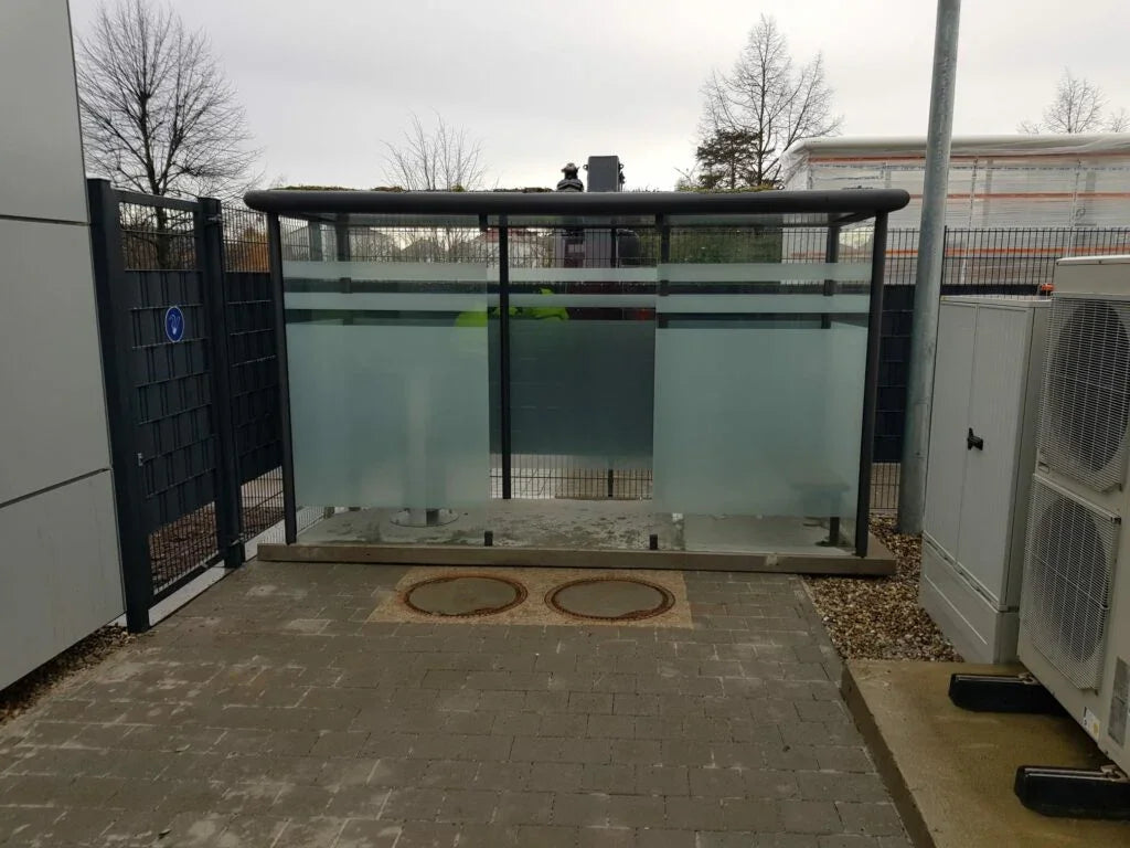 Moderne Raucherkabine aus Glas und Metall für den Außenbereich, auf Pflastersteinen.