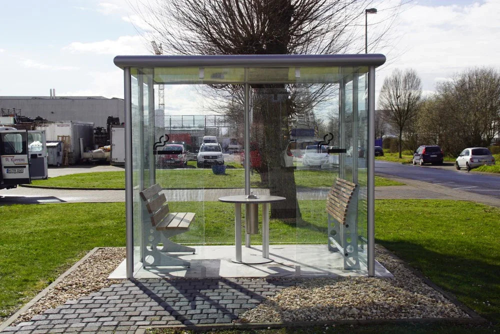 Raucherkabine aus Glas und Metall mit Sitzbänken und Tisch im Außenbereich