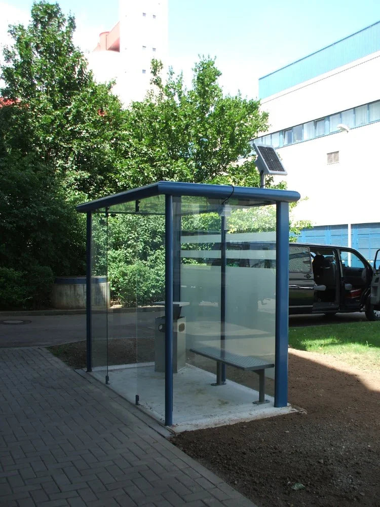 Freistehende Raucherkabine aus Glas und Metall im Außenbereich mit Sitzbank