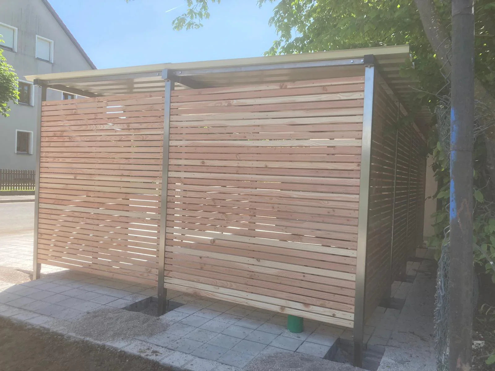 Modernes Fahrrad- und Mülltonnenhaus aus Metall und Holz im Garten auf Pflasterboden.