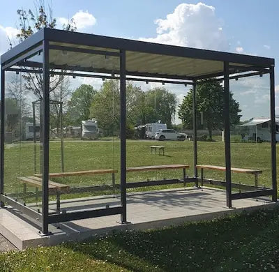 Moderne Wartehalle aus Metall und Glas mit Sitzbänken, auf Wiese bei Tageslicht