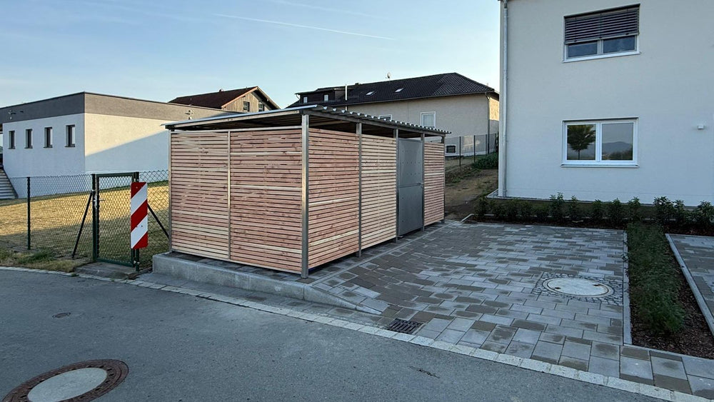 Fahrradgarage mit Holzverkleidung und abschließbarer Metalltür auf Betonfundament neben einem Wohnhaus