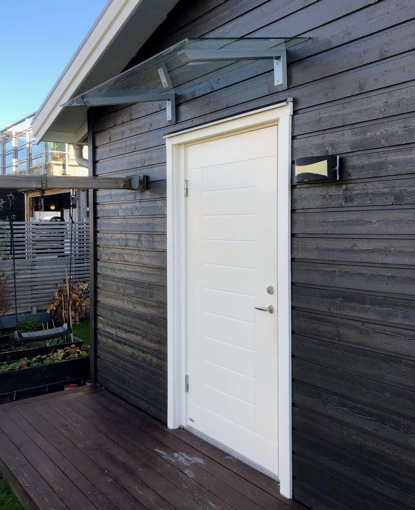 Vordach aus Klarglas mit silbernen Konsolen über weißer Haustür an schwarzer Holzfassade