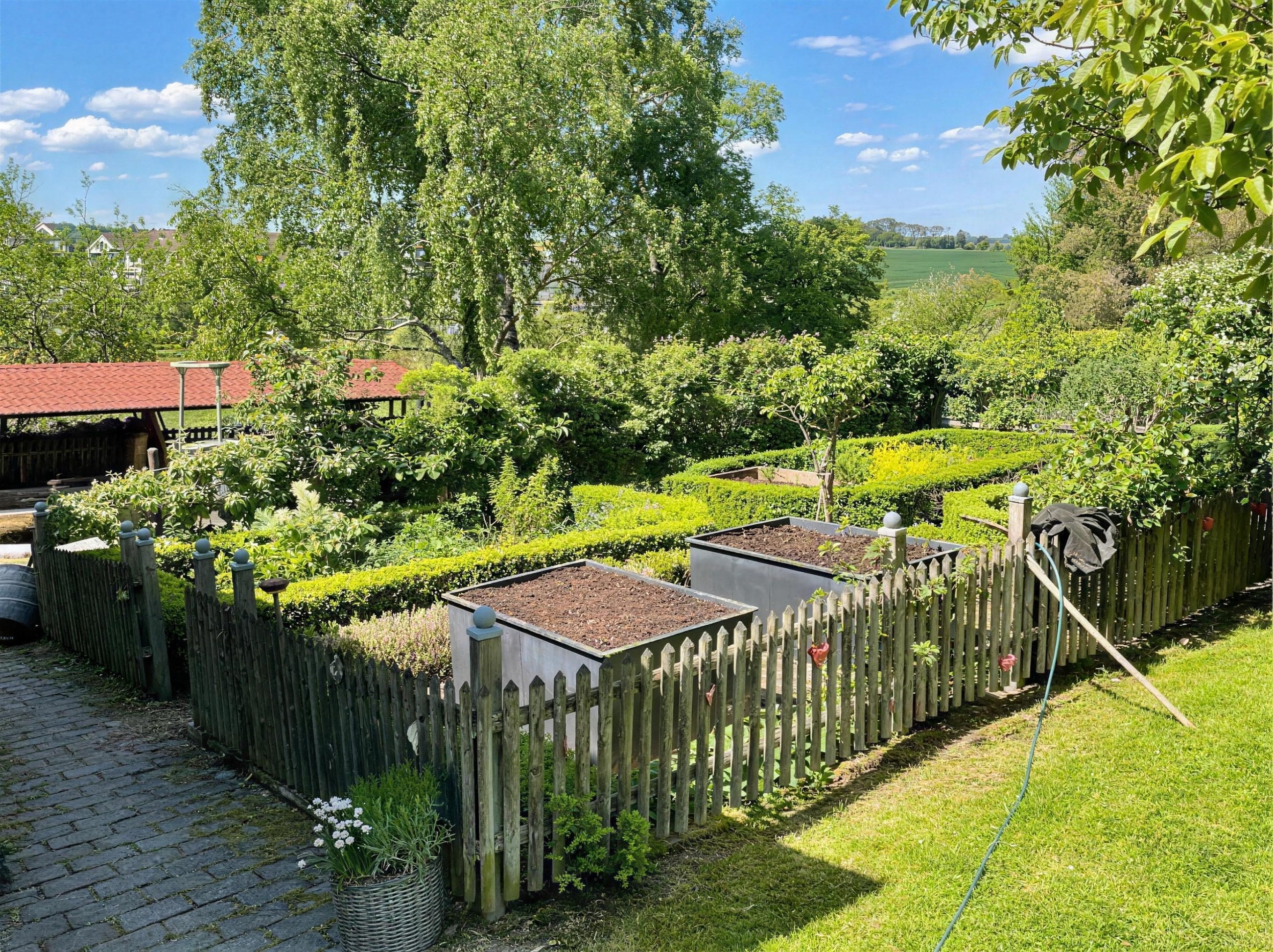 Kundenfoto: zwei Cortenstahl Hochbeete nach Maß im Bauerngarten hinter Staketenzaun mit Buchsbaumhecke