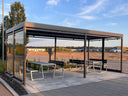 Moderne Metallüberdachung mit seitlichen Glaswänden, Sitzbänken und Tisch auf gepflastertem Platz
